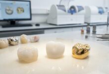 A photorealistic close-up of three distinct dental crowns—a translucent ceramic, a solid white zirconia, and a polished gold one—set against the blurred background of a modern dental lab.