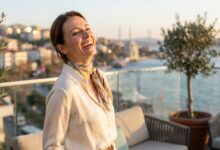 A radiant woman with a flawless, bright white smile, standing on a balcony overlooking a blurred coastal Turkish city at sunset, symbolizing a successful smile makeover.