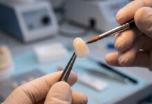 A close-up, photorealistic image of a dental technician's hands carefully crafting a single porcelain veneer with a small brush in a modern, clean laboratory setting.