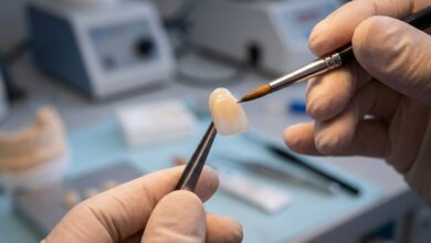 A close-up, photorealistic image of a dental technician's hands carefully crafting a single porcelain veneer with a small brush in a modern, clean laboratory setting.