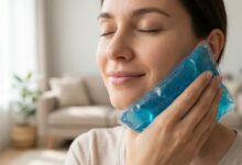 A close-up, photorealistic photo showing a person with a calm expression holding a soft gel ice pack against their jaw, illustrating post-operative self-care in a peaceful home setting.