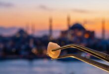 A close-up, photorealistic image of a perfect white zirconia dental crown held by tweezers, with the sun setting over a heavily blurred background of the Istanbul, Turkey skyline and the Bosphorus.