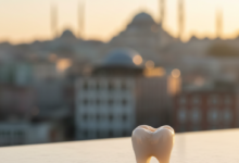 A close-up, photorealistic image of a perfectly crafted, white dental crown on a clean surface. The background is a blurred, soft-focus image of Istanbul, Turkey, showing faint architectural outlines of a mosque or city buildings.