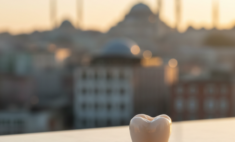 A close-up, photorealistic image of a perfectly crafted, white dental crown on a clean surface. The background is a blurred, soft-focus image of Istanbul, Turkey, showing faint architectural outlines of a mosque or city buildings.