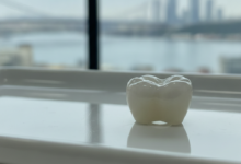 A photorealistic close-up of a gleaming, perfectly shaped dental crown, appearing on a sterile, light-reflecting surface in a clean, modern dental environment. A very soft, out-of-focus background shows blurred lights and shapes suggestive of a Turkish city skyline.