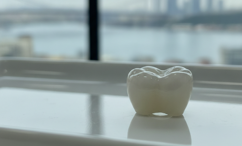 A photorealistic close-up of a gleaming, perfectly shaped dental crown, appearing on a sterile, light-reflecting surface in a clean, modern dental environment. A very soft, out-of-focus background shows blurred lights and shapes suggestive of a Turkish city skyline.