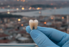 Close-up of a perfectly crafted, natural-looking dental crown on a gloved fingertip or dental tray. The background shows a beautifully blurred, warm-toned Turkish city skyline, hinting at a modern dental tourism destination.
