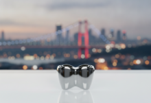 A close-up, photorealistic view of a gleaming, perfectly crafted white dental crown, positioned centrally with a shallow depth of field. In the blurred background, a modern Turkish city skyline featuring a bridge and city lights is discernible, suggesting Istanbul. The image highlights precision and advanced dental care.
