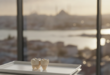 A photorealistic image of a modern dental clinic interior. In the foreground, a single, high-quality, natural-looking dental crown is displayed on a sterile white tray, in sharp focus. The background shows a softly blurred, warm-toned cityscape of Istanbul through a large window, suggesting Turkey as a premier destination for dental care.