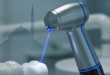 A photorealistic image of a futuristic dental laser handpiece emitting a precise light beam towards a generic tooth or gum, rendered in ultra-detail with a shallow depth of field. The background is a softly blurred modern dental clinic, with a very subtle, distant outline of Turkish architecture. No visible text.